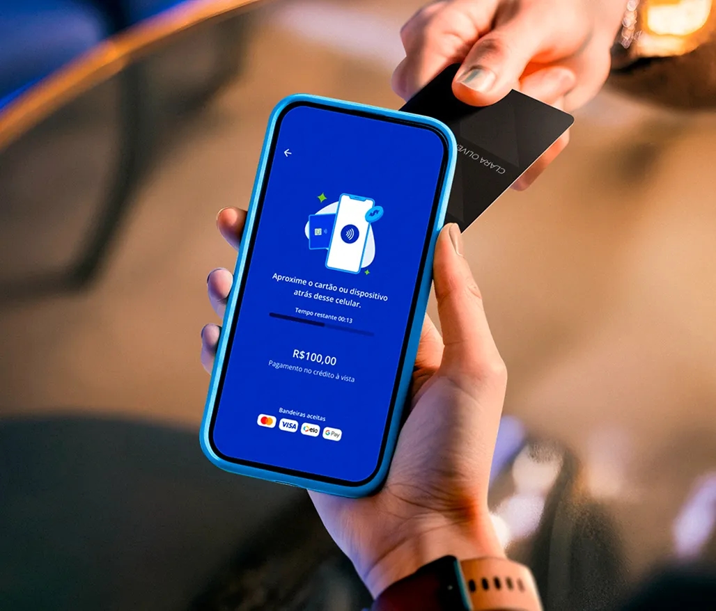 Pessoa segurando um celular com a tela exibindo a interface do Asaas com a mensagem: "Aproxime o cartão ou dispositivo atrás desse celular". Outra pessoa aproxima um cartão para efetuar o pagamento.