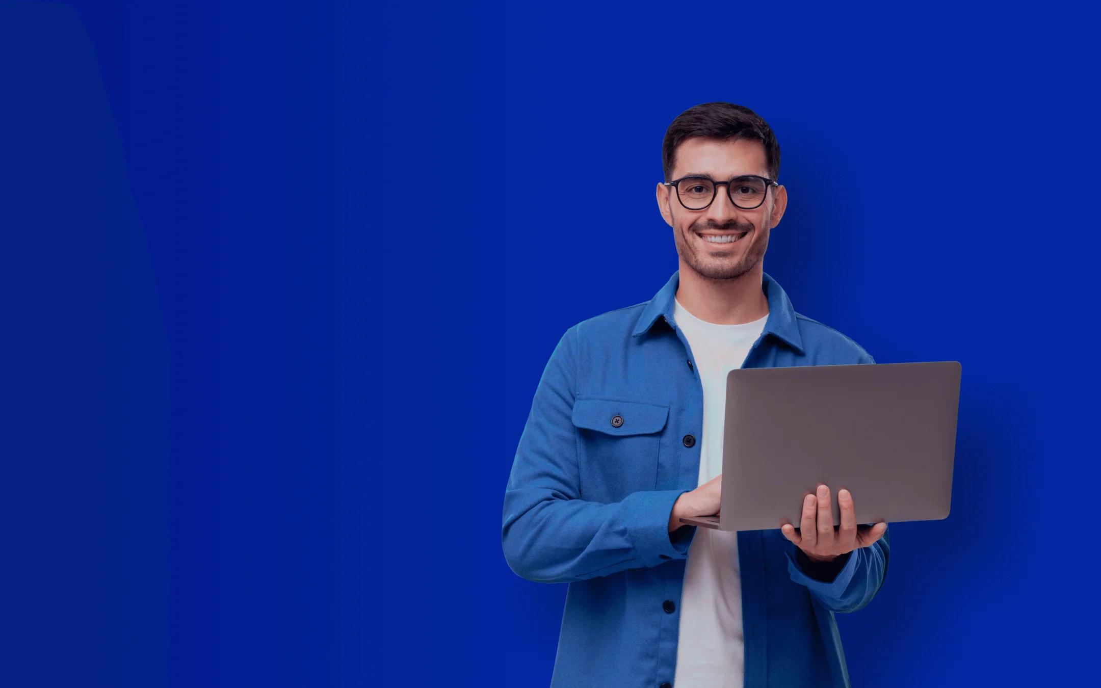 Homem branco de cabelo escuro e barba rala usando óculos de armação preta sorri para a câmera. Ele está de pé com uma camisa azul clara aberta e usa camiseta branca. Ele segura um notebook com as mãos.