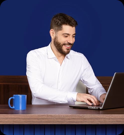 Evaristo Costa veste uma camisa social branca e está sentado em uma escrivaninha digitando em um notebook.