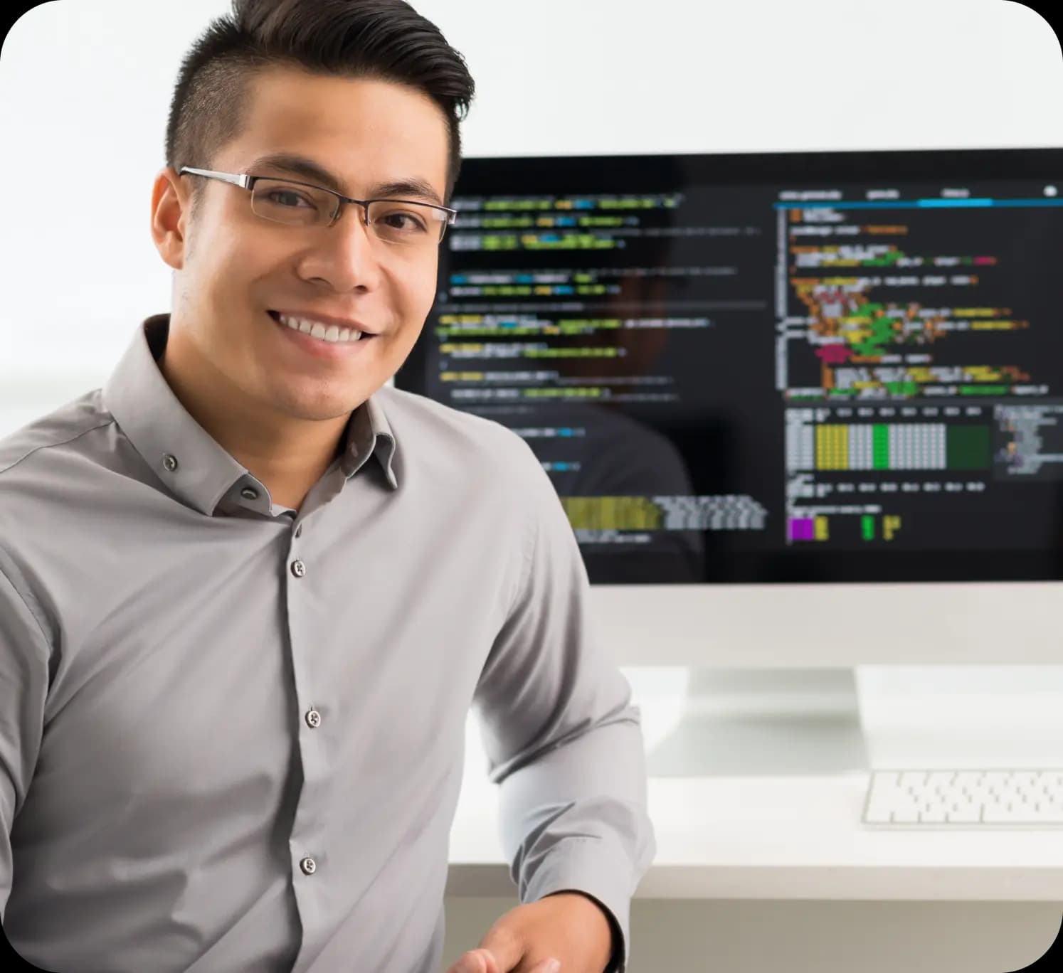 Profissional de tecnologia da informação sorri enquanto olha para frente. Ele tem cabelos escuros e curtos, pele clara, e veste uma camiseta social cinza de botão. Ao fundo, há um computador do tipo “all-in-one” mostrando um código de programação da API de pagamentos.