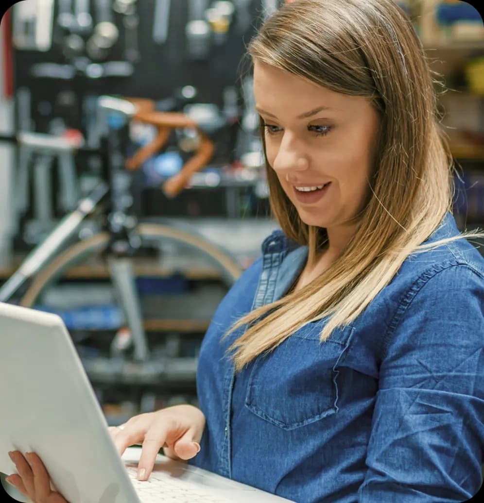 Empreendedora mexe no computador. Ela tem cabelos lisos, compridos e loiros, e veste uma camiseta de botão em tecido jeans. Ao fundo, em desfoque, está uma loja de materiais, com uma bicicleta próxima.