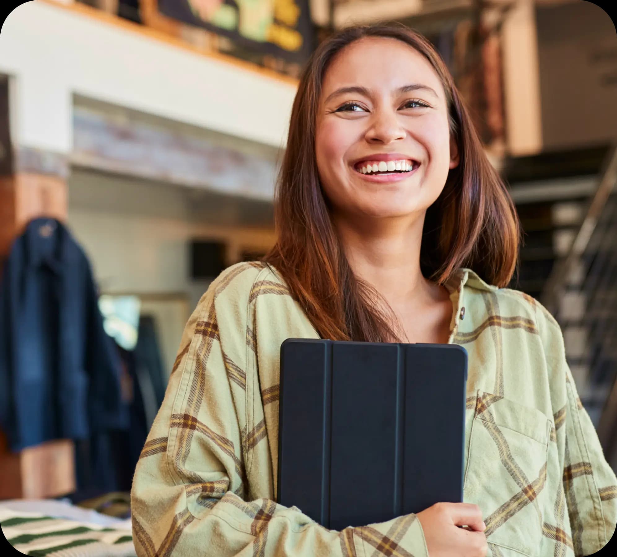 Empreendedora sorri, enquanto olha para sua direita. Ela tem pele clara e cabelos lisos, com tons castanhos e acobreados. Também veste uma camisa de botão xadrez, nas cores verde e marrom. Ela segura um tablet com a mão direita, junto ao corpo.