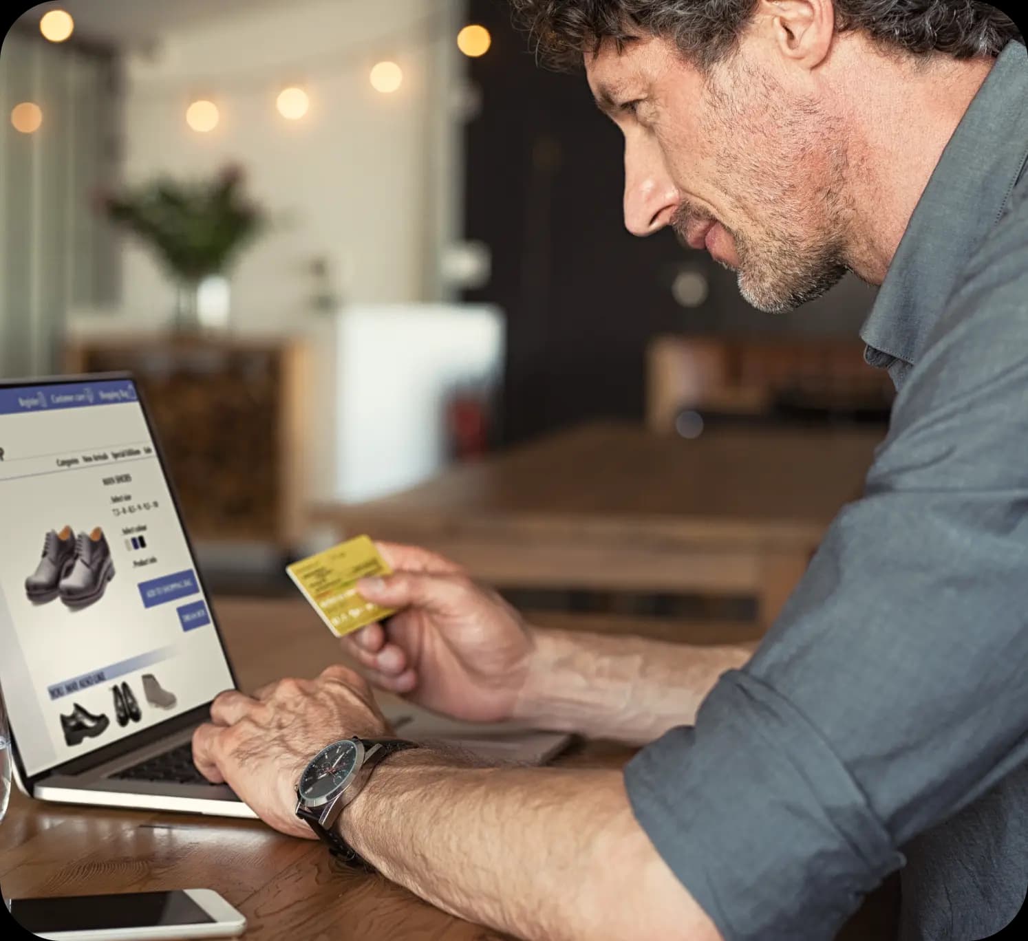 Homem segura cartão de crédito e olha para a tela de um notebook. Essa tela mostra o site de uma loja online, onde está exposto um sapato social. O homem tem a pele clara, cabelo curto e barba por fazer. Ele veste uma camiseta social cinza e relógio de pulso. Ao fundo, o cenário lembra o de uma residência.