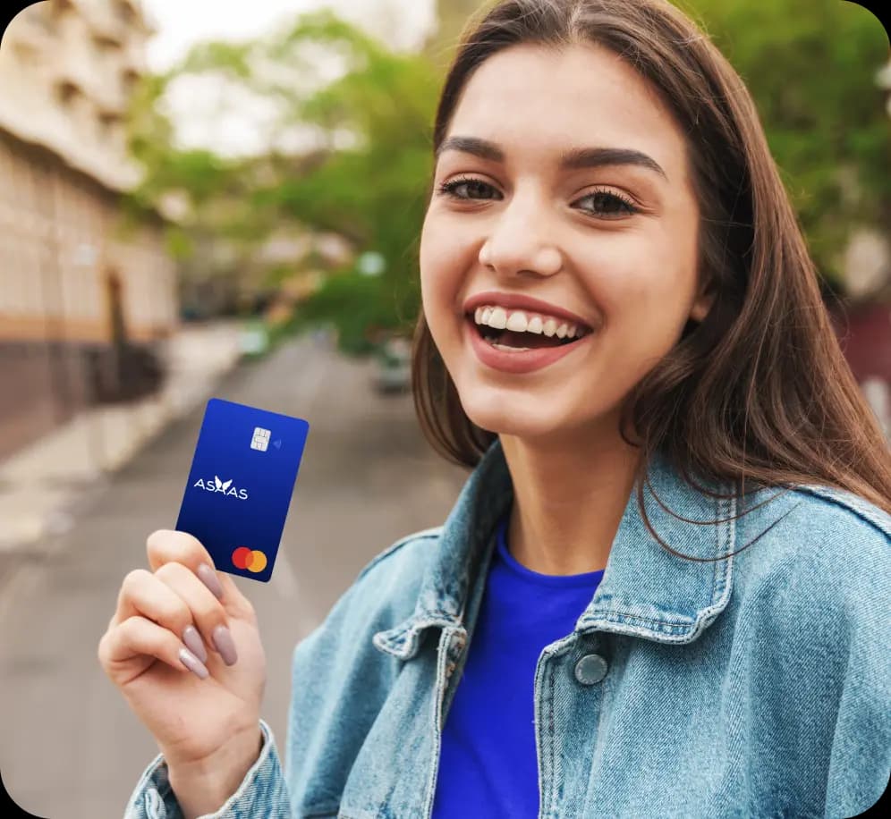 Empreendedora sorri para a câmera enquanto segura o Cartão Asaas. Ela tem cabelos longos, lisos e castanhos, pele clara, e veste uma camiseta azul e jaqueta jeans.