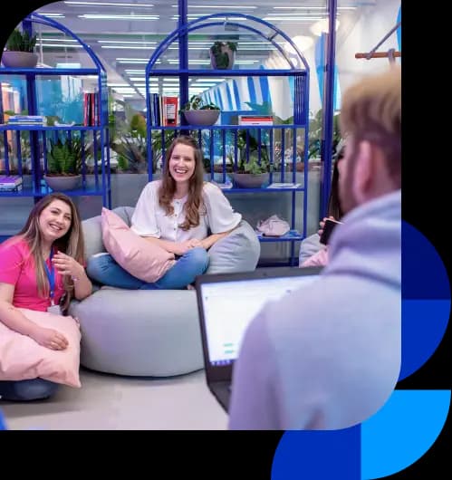 Colaboradores do Asaas conversam na área criativa. Há duas mulheres sorrindo, uma está sentada em um puff e a outra de joelhos no chão, com uma almofada no colo. No canto inferior direito, há um homem de costas, em desfoque. Ao fundo, estão duas estantes coloridas em azul e algumas plantas.