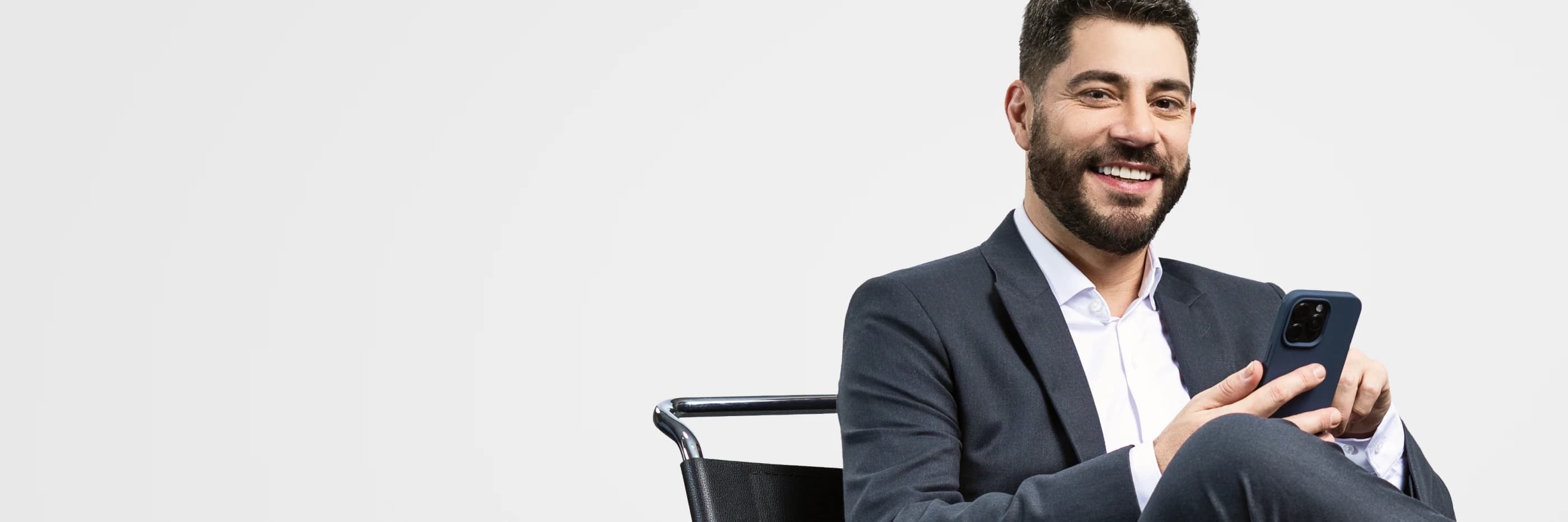 Evaristo Costa veste um terno e está sentado em uma cadeira com um celular na mão.