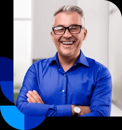 Homem branco de cabelos grisalhos, com os braços cruzados e sorrindo para a câmera. Ele está vestindo uma camisa social azul, óculos de armação escura e um relógio em seu pulso direito.