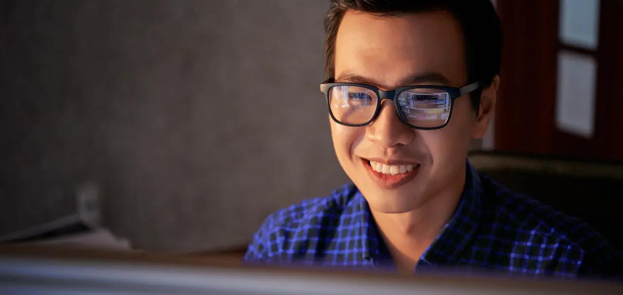 Desenvolvedor sorri para a tela do computador enquanto usa a API de pagamentos do Asaas. Ele tem a pele clara, cabelos curtos e castanhos, e usa óculos de grau com armação preta. Além disso, veste uma camiseta xadrez na cor azul. Ao fundo, há uma parede cinza e uma janela.