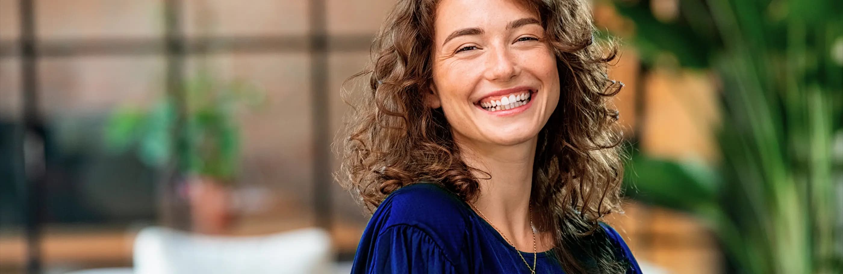 Mulher branca de cabelos castanho claro e cacheado sorri para a foto com ar de satisfação. Ela veste uma camisa azul escuto e um colar dourado.