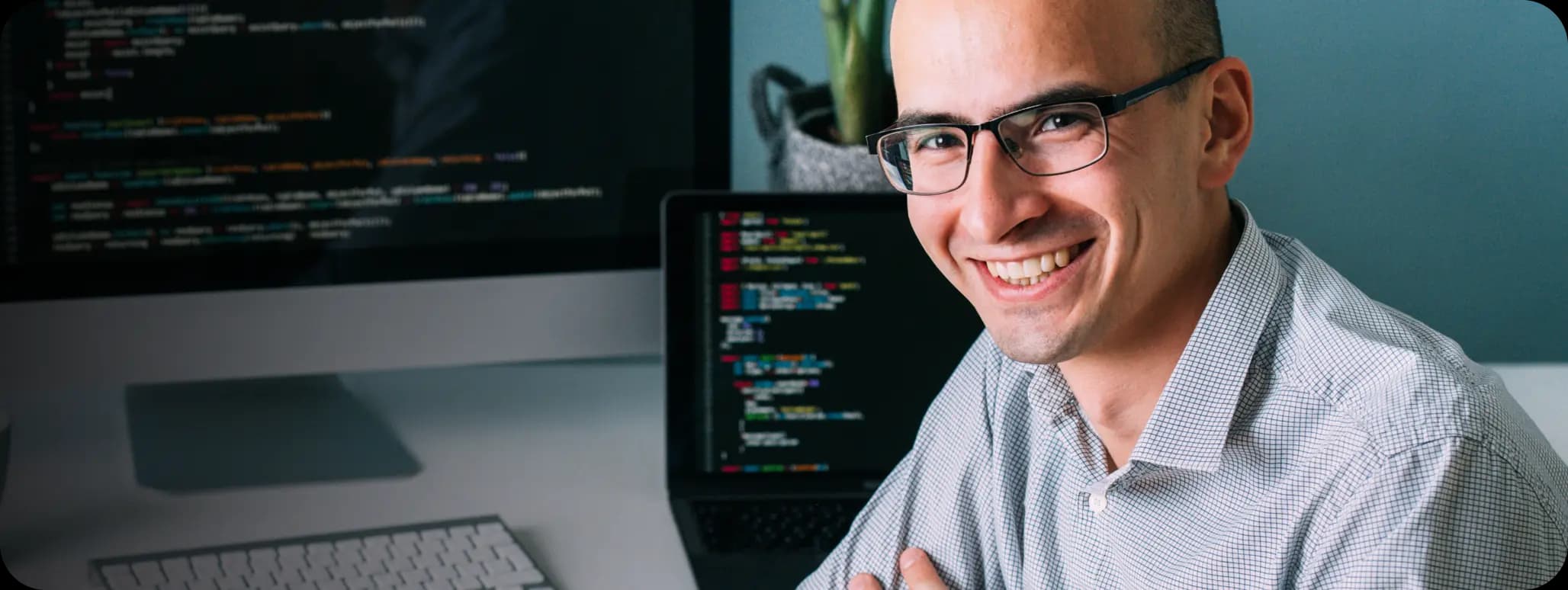 Homem branco, careca e que usa óculos de grau sorri para a foto com os braços cruzados apoiados em uma mesa. Veste uma camisa social com textura quadriculada azul clara.