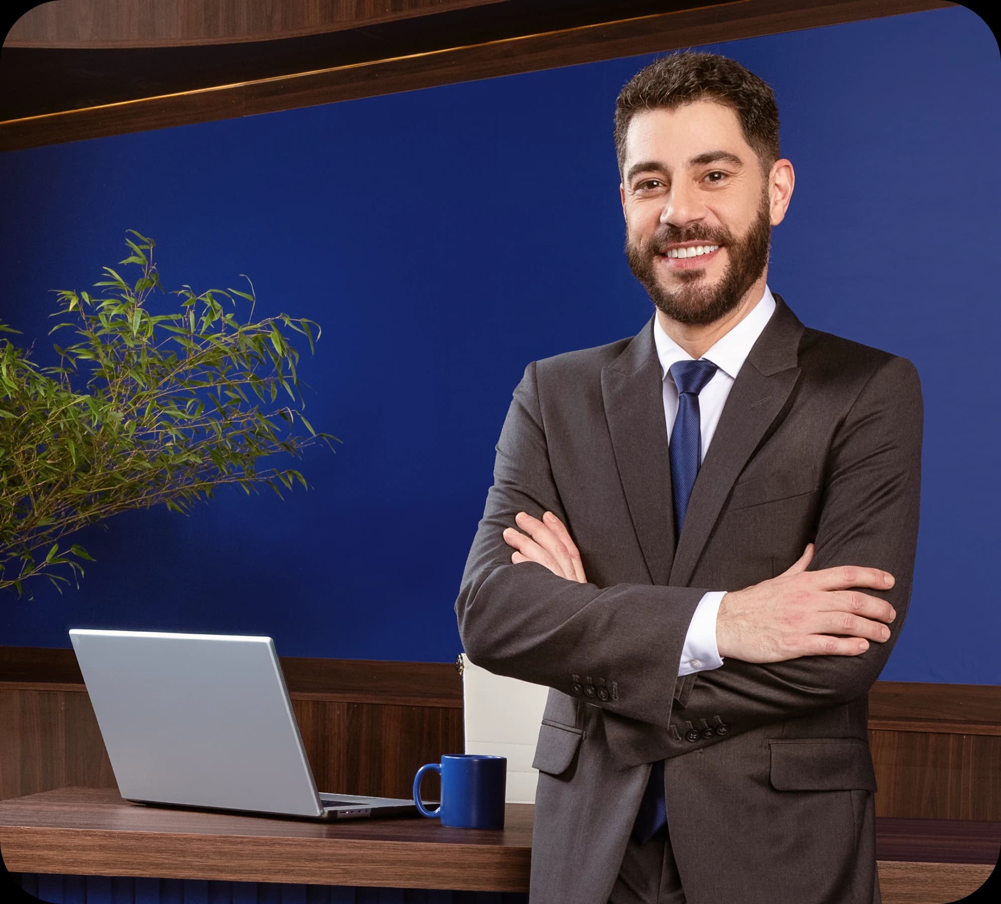 Evaristo Costa veste um terno e está de braços cruzados e sorrindo apoiado em uma mesa que tem um notebook e uma xícara em cima.